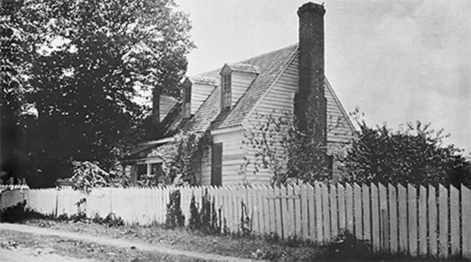 Black and white photo of wooden, two-storey house with brick chimney, second-floor dormers, and a white picket fence in front. 